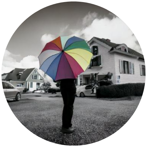 Person holding a colorful umbrella in front of a suburban house with cars parked, under a cloudy sky